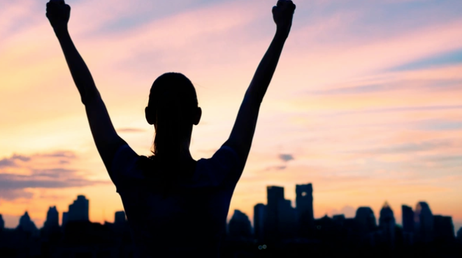Orange and pink sunset in the background. Forefront is all black with a city outline and the shape of a woman with her hands raised in the air triumphantly.