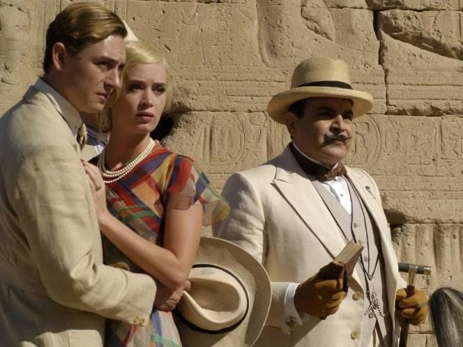 Simon Doyle (JJ Feild), Linnet Ridgeway (a blond Emily Blunt) and Hercule Poirot (David Suchet in a white suit) stand in front of the pyramids looking at something offscreen.