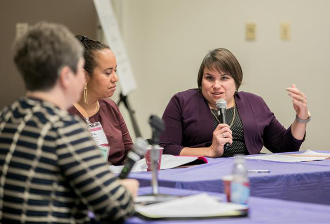 Karen sits at a conference table, smartly dressed, speaking into a microphone. A woman next to her listens attentively.