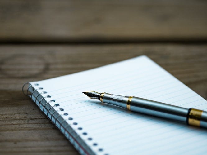 A blank spiral-bound notebook and a silver and gold fountain pen resting on a greyish-brown wooden table.