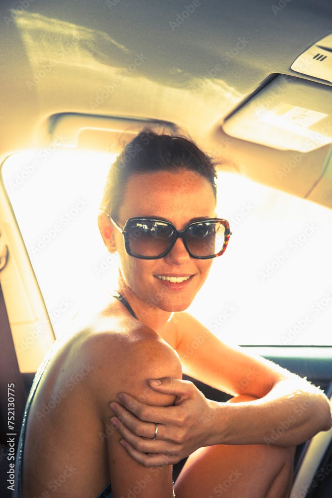 Beautiful   woman sitting in a car with big sun glasses and a black bikini on. the sun is shining on her, and her hair is pulled back into a pony tail.