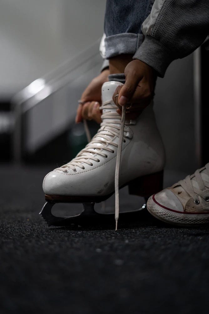 Close up of a white ice skate and two brown hands tying the laces. Background is blurred and grey and a dingy white converse sneaker peaks out on the lower right hand corner.