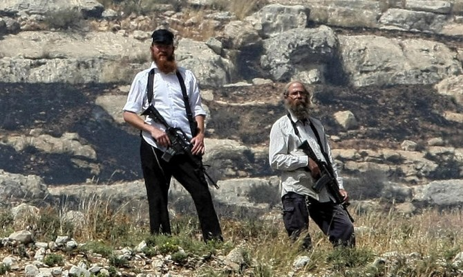 A imagem mostra dois colonos israelenses posando para a câmera ao ar livre, próximos de um assentamento ilegal na Cisjordânia. Eles estão em um terreno rochoso com grama e plantas ao seu redor. Esses homens carregam consigo armas de fogo de grande porte.