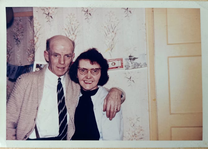 A balding white man in his sixties has his arm around a dark haired woman of the same age who is wearing glasses. They stand against a wall by a door, there is a string of Christmas cards displayed behind them.