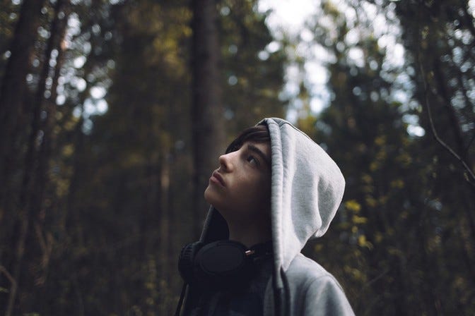 Middle school male looking up into the trees in nature with headphones around his neck and his hood up.