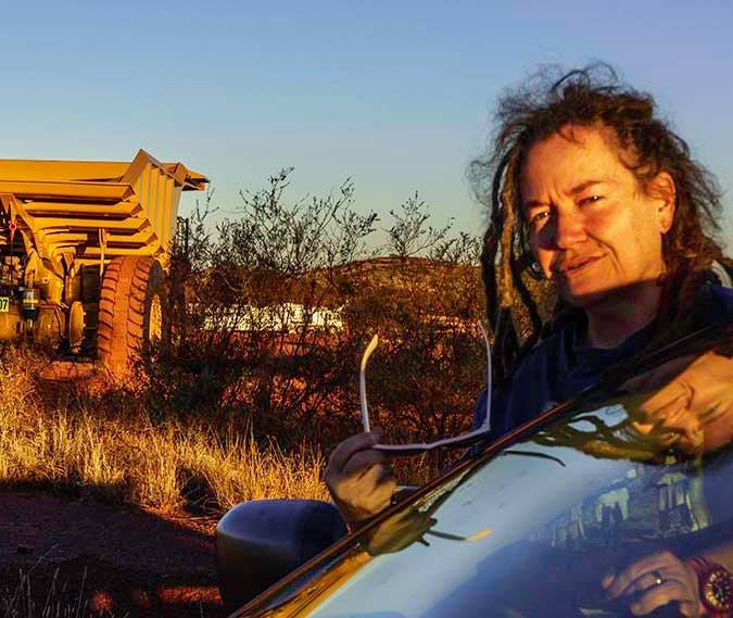 A tired woman with dreadlocks and messy hair, sunglasses in hand, frowns near a car windscreen. A supersized mining truck and dried plants in the background.