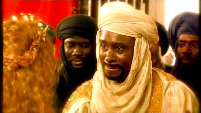 A Black man wearing a white turban and an ornate tunic faces a redheaded woman. Behind him, stand three other Black men wearing black turbans.