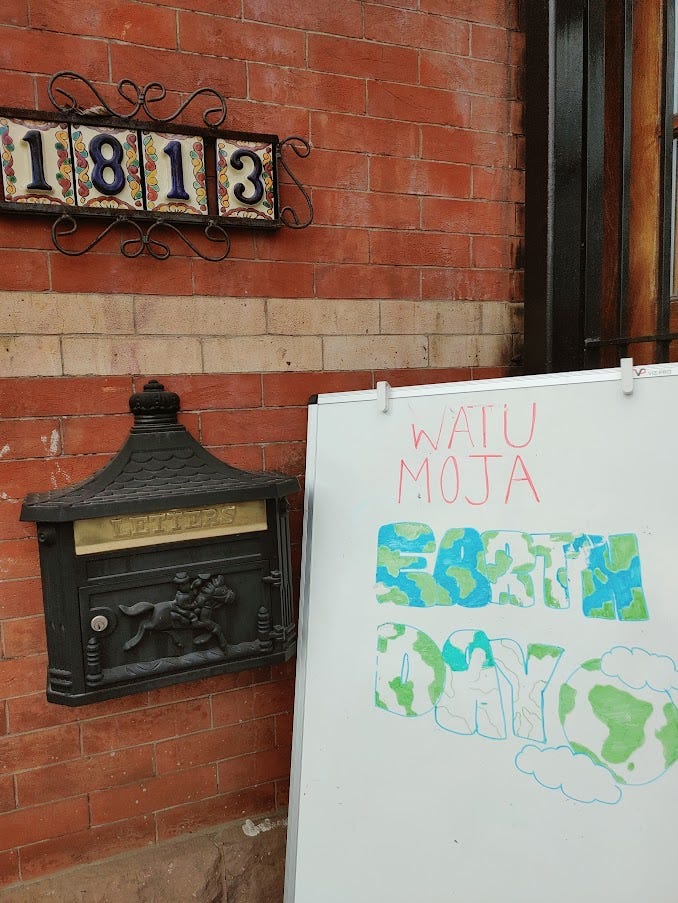A whiteboard sign leaning up against a brick wall. The writing on the board says “WATU MOJA: EARTH DAY.”
