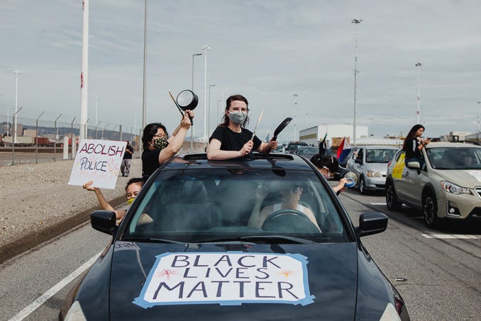 A group of people lean out of the windows of a car, holding signs that say “abolish the police” and “Black Lives Matter.”