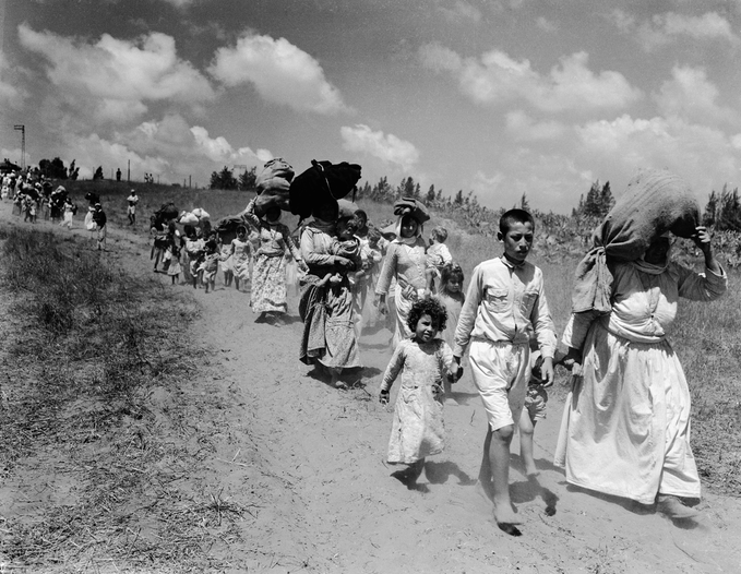 A imagem é uma fotografia histórica em preto e branco que retrata uma fila de famílias palestinas durante a Nakba. Eles caminham por uma estrada de terra, foram expulsos das suas casas e terras num período caracterizado pela deslocação do povo palestino.