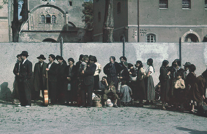 Fotografia colorida onde retrata ciganos sendo detidos para deportação, pelo governo alemão, em 22 de maio de 1940. Nessa foto, numa rua, próximos de um muro, existe uma fila e aglomerado de pessoas ciganas: homens, mulheres, crianças.