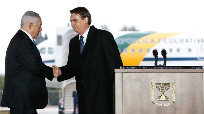 Dois homens vestidos de terno preto, identificados como o primeiro-ministro israelense, Benjamin Netanyahu, à esquerda, e o presidente do Brasil, Jair Bolsonaro, à direita, apertando as mãos na pista.