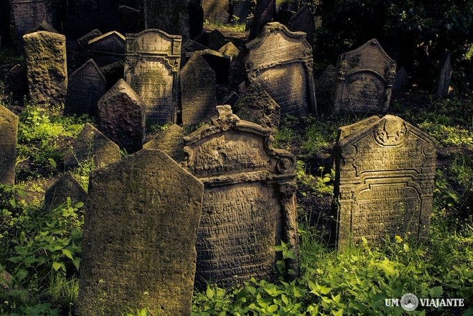 A fotografia mostra um grupo de lápides com inscrições do Novo Cemitério Judaico, em Praga. Uma das pedras traz o texto “um aviajante”.