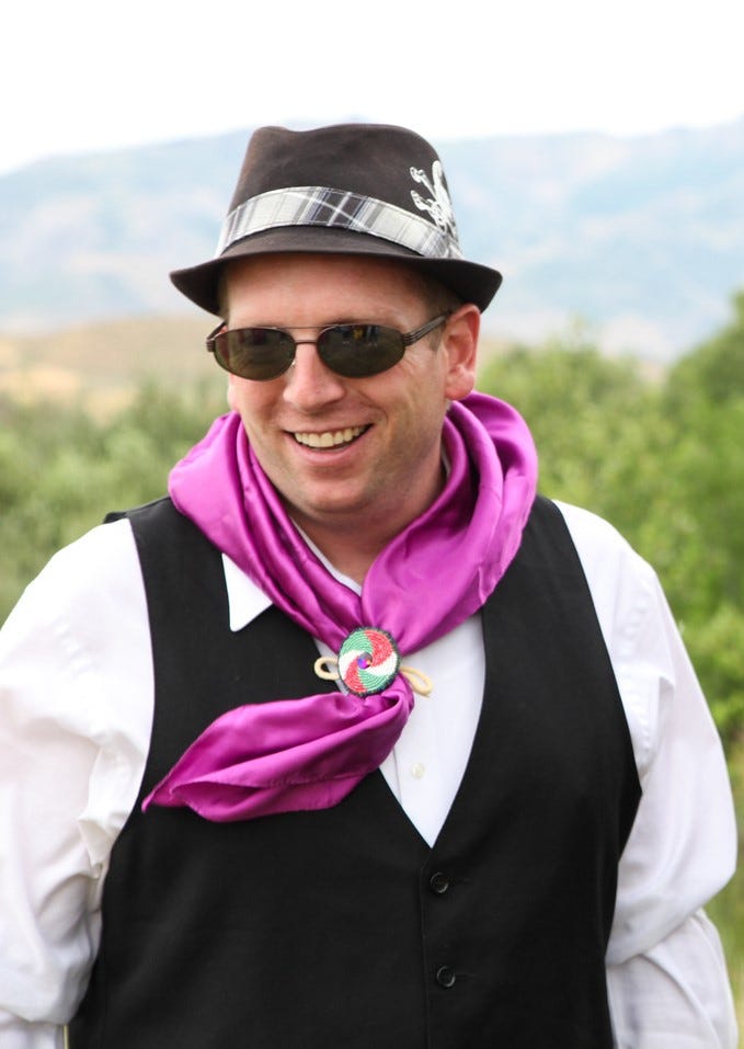 Shsohone man wearing a hat and traditional scarf smiling.