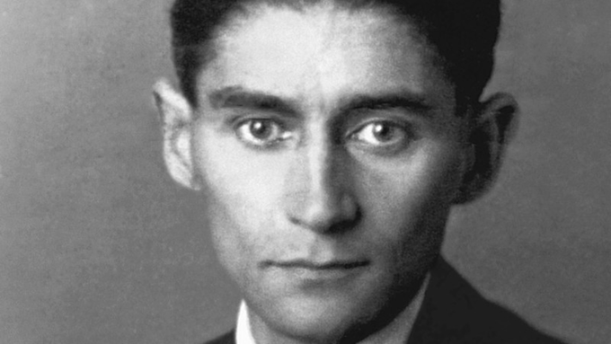 A imagem apresenta o retrato de Franz Kafka com uma cara séria. A foto em preto e branco concentra-se nas características faciais do homem, como sobrancelhas, testa, queixo e mandíbula. Ele está dentro de casa e parece estar vestindo um terno.