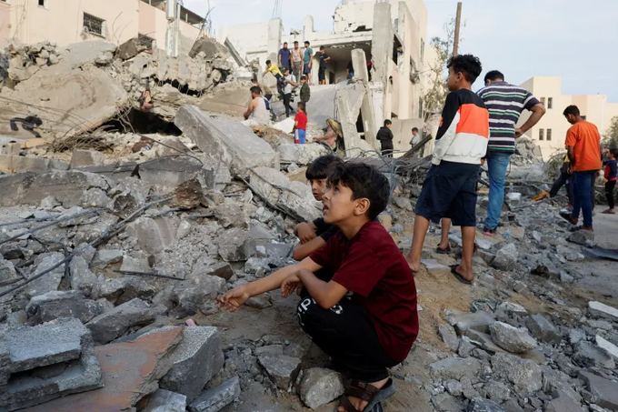 A imagem mostra um grupo de palestinos, incluindo crianças, em meio a um monte de escombros de um edifício em Gaza bombardeado por Israel. Eles estão vestindo diferentes tipos de roupas e calçados.