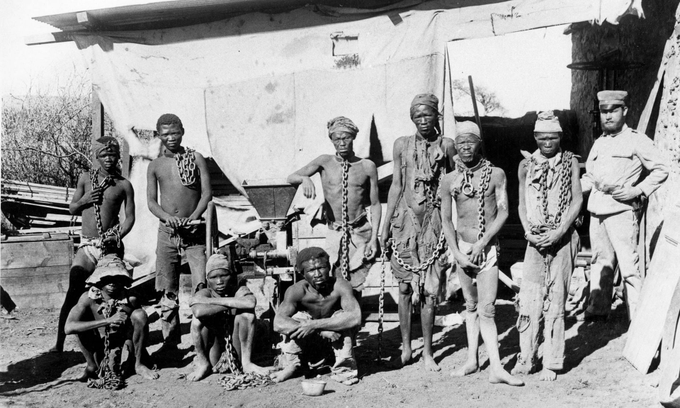 Uma fotografia em preto e branco de prisioneiros Herero e Nama acorrentados durante o genocídio, num campo de concentração.