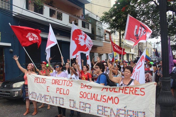 Fotografia de mulheres feministas do Movimento Olga Benário segurando bandeiras com o rosto Benário e da UJC. Mulheres da linha de frente da manifestação seguram um tecido branco longo que diz “Pelos direitos das mulheres e pelo socialismo”.