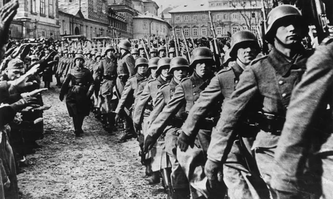 A imagem mostra um grupo de soldados marchando. Eles estão vestindo uniformes militares e a foto é em preto e branco. Os soldados estão lá fora marchando em grupo e há um prédio ao fundo.