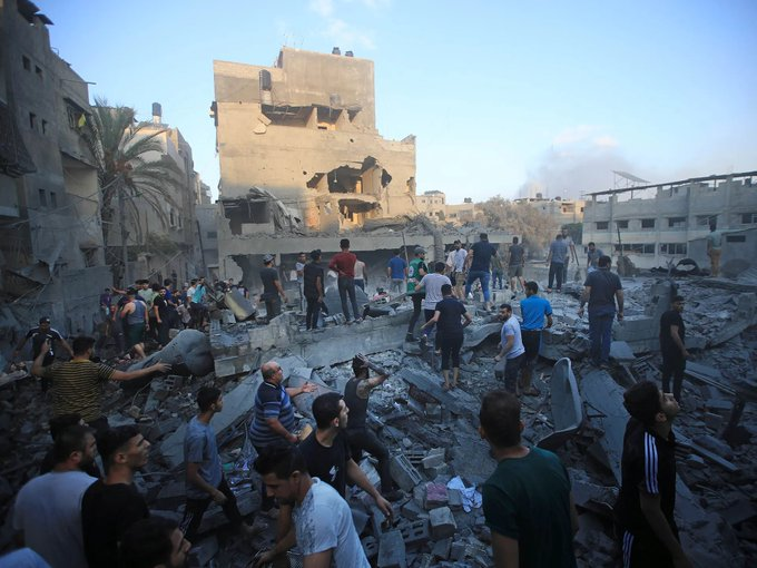 A fotografia mostra um grupo de muitos palestinos, na maioria homens, em uma rua com prédios e edifícios destruídos após serem bombardeados por Israel, com muitos destroços e escombros.