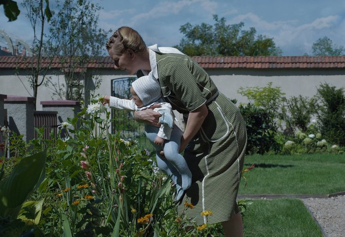 A imagem mostra Hedwig Höss segurando um bebê ao ar livre em um jardim durante o verão, se curvado para observar suas plantas. Os arredores incluem árvores, grama, flores e um céu claro.