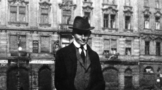 A imagem mostra um Franz Kafka vestindo terno e chapéu em um ambiente urbano. É uma fotografia em preto e branco, e Kafka está vestido de cavalheiro, usando chapéu de feltro.