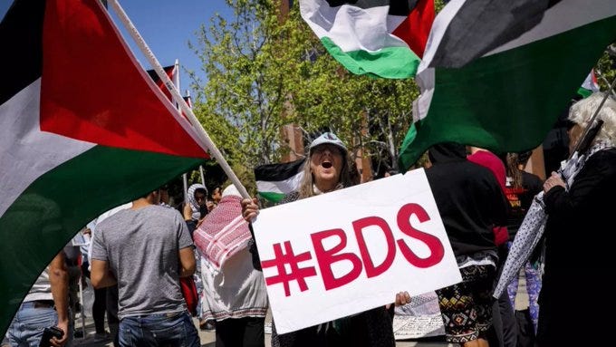 A imagem captura uma manifestação ou protesto numa rua da cidade, com participantes demonstrando apoio ao movimento #BDS sob um céu azul brilhante. Numerosas bandeiras palestinas estão sendo erguidas pelos manifestantes, em cores que incluem verde, branco, preto e vermelho. Pelo menos nove indivíduos estão visíveis na cena. Uma placa com a hashtag “#BDS”.