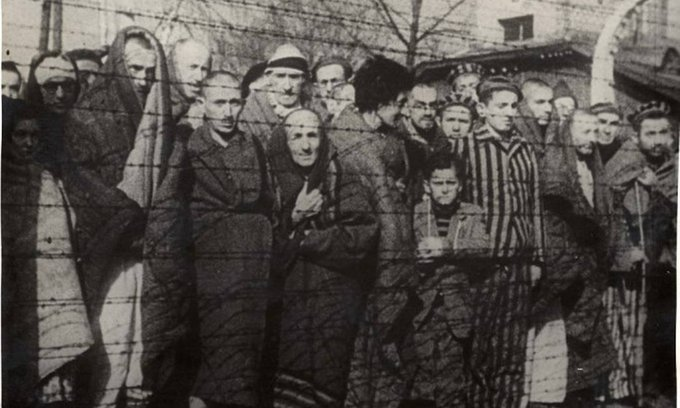 Fotografia em preto e branco de sobreviventes do Holocausto atrás de cerca de arame farpado no dia da liberação de Auschwitz.