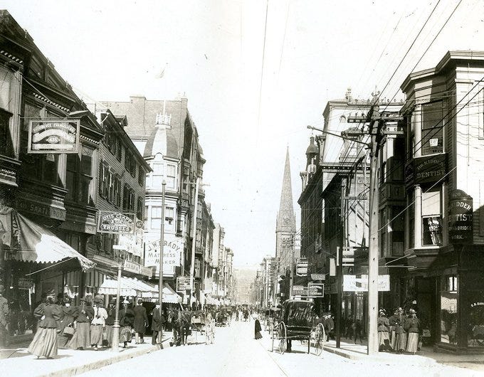 A black and white photo of a busy street crowded with people including women with ponytails and puffed sleeves; a horse-drawn cart and a church steeple in the distance.