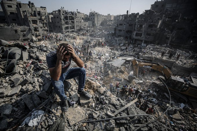 A imagem mostra um homem palestino sentado sobre uma pilha de destroços e escombros. Ele está vestindo jeans e parece estar ao ar livre sob um céu claro. O entorno apresenta uma situação de plena destruição de edifícios palestinos, na Faixa de Gaza, como resultado dos bombardeamentos de Israel contra habitantes de Gaza.