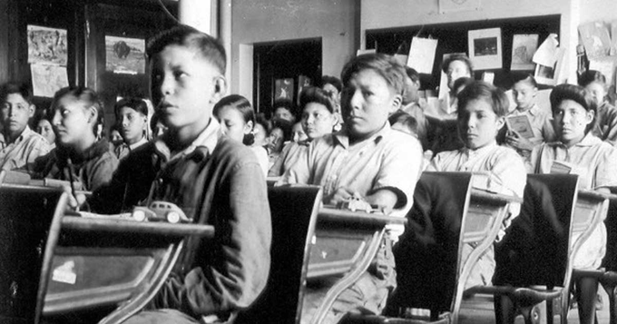 Fotografia em preto e branco de uma sala de aula de uma escola residencial para indígenas Old Sun, no Canadá. Crianças indígenas sentadas às carteiras, como alunos.