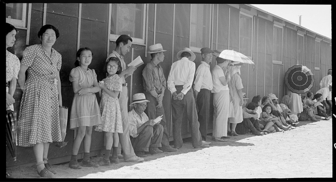 A imagem é uma foto em preto e branco que mostra um grupo de pessoas japonesas e nipo-americanas do lado de fora de uma moradia, dentro de um campo de concentração nos EUA, na década de 1940. Os indivíduos estão vestidos com vários tipos de roupas e calçados.