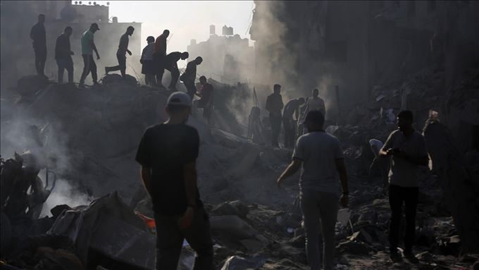 Fotografia colorida de escombros na Faixa de Gaza, onde um grupos de palestinos estão ao redor observando a situação e tentando erguer os destroços para socorrer as vítimas.