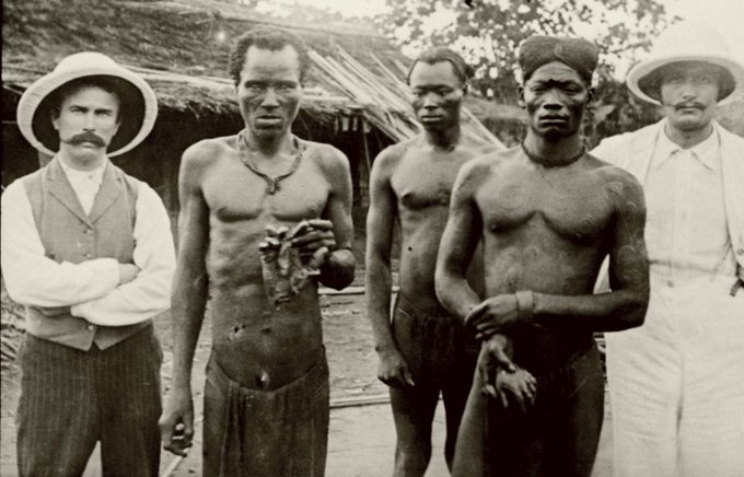 A imagem mostra um grupo de colonos belgas posando para uma foto ao ar livre, num cenário de violência contra homens congoleses, que estão algemados e com ferimentos. A foto está em preto e branco.