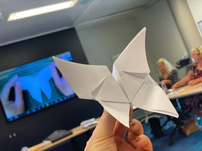 A hand holding a white paper origami butterfly. Some people and a presentation screen in the background.