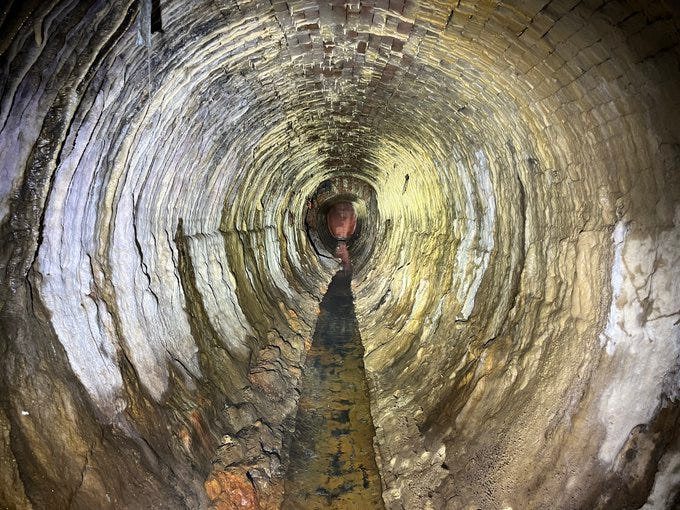 brick lined 152 year old sewer. brick is brown and rounded but intact. mineral deposits line the edges and murky water flows gently down the center of the sewer invert towards the darkness.