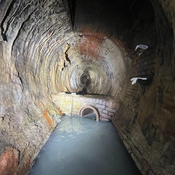 brick-lined sewer as seen from standing inside. 149 years old a flashlight illuminates the brick. a walled regulator is visible several paces in front of the photographer standing in murky dirty water in the middle of the invert.