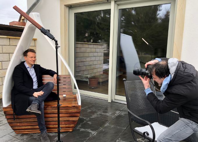 A man sits in a tear-drop-shaped chair made of a wind turbine rotor blade and wood. A man is taking his photo.