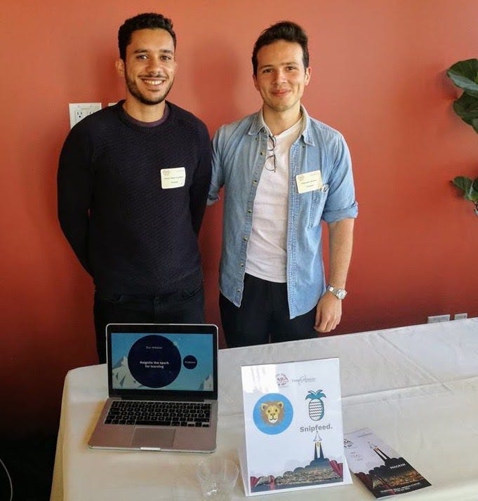 Two young men pose behind a white table.