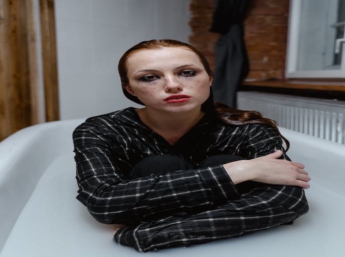 An image depicting a woman sitting in a white bathtub, wearing a black and white plaid shirt, her expression conveying a mix of confusion, frustration, and vulnerability, reflecting the impact of identity theft and online insecurity.
