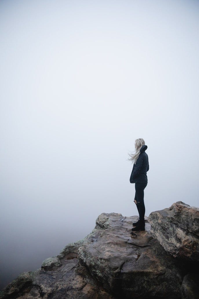 A slender lady stands on the hill tops with hands in pocket and admires the hilly surrounding and the fresh air.