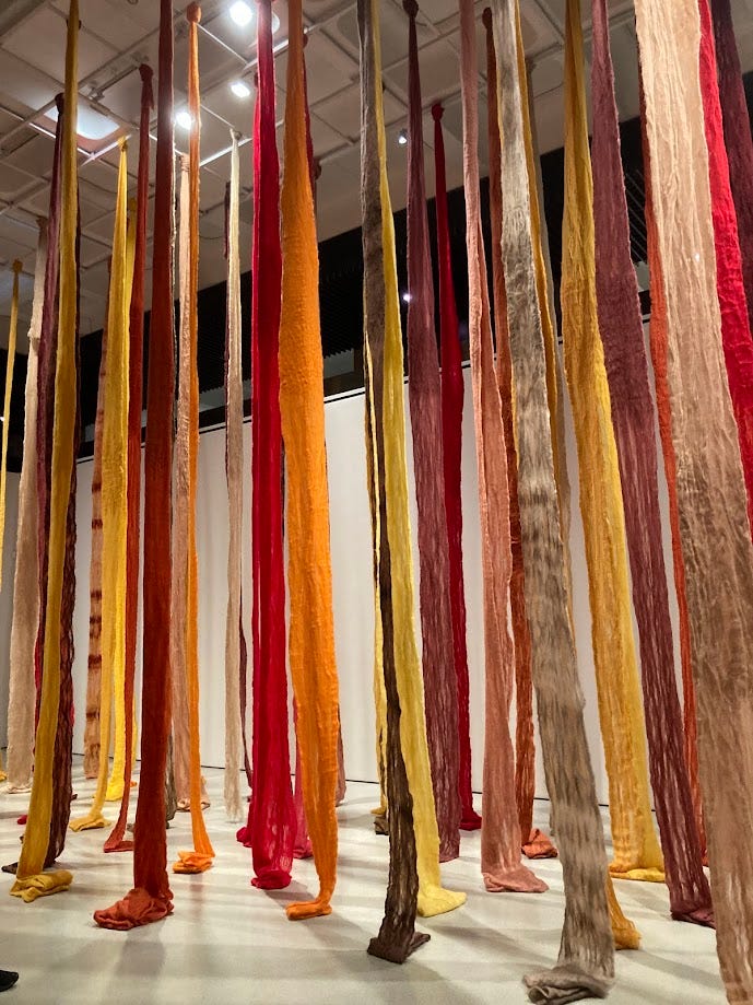 Long strands of cloth in bright oranges, yellows and reds dangle from the ceiling of an art gallery.