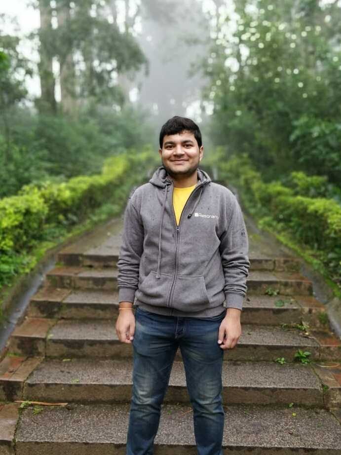 A person wearing a gray hoodie and jeans stands outside on concrete stairs, surrounded by greenery. He smiles at the camera.