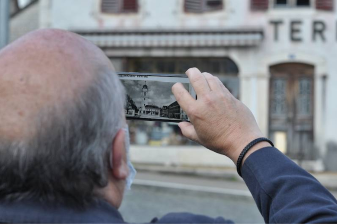 An old man looking at the front of a building through a smartphone application. On the phone, the building is in black and white