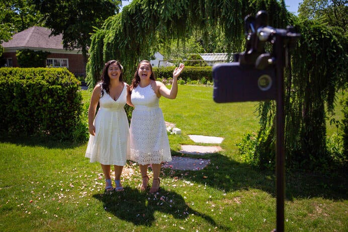 Two brides wave to Zoom wedding guests on an iPhone at a 2020 Covid Wedding.