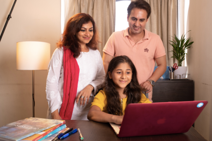 Smiling mom, dad and daughter looking into the laptop, kept on wooden table with a plant and a floor lamp in the background.
