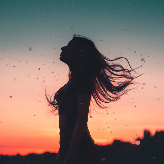 Silhouette of a woman standing against the backdrop of a colorful sunrise sky, with wind blowing through her hair and the environment. Image created in Canva, using a PIxabay image by jakovzadro1