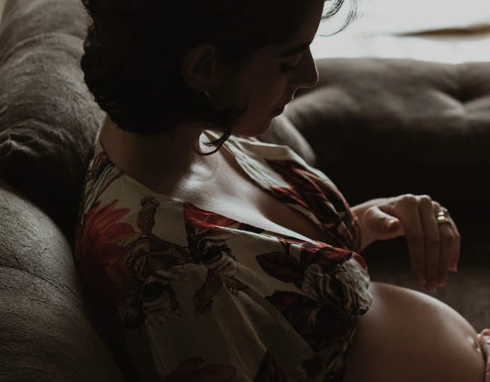 Pregnant mother holds baby bump in a dimly lit room while seated on a couch.