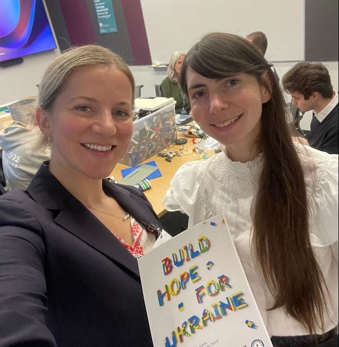 Two women holding a “build hope for Ukraine” sign with LEGO bricks in the back