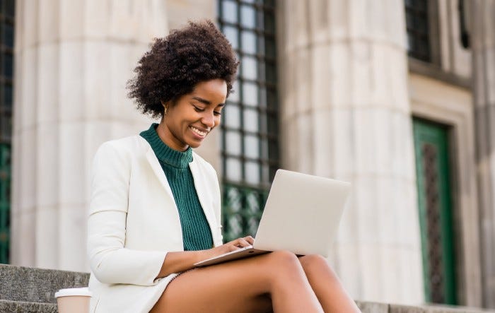 Business woman using her laptop while sitting on stairs of a college building outdoors.